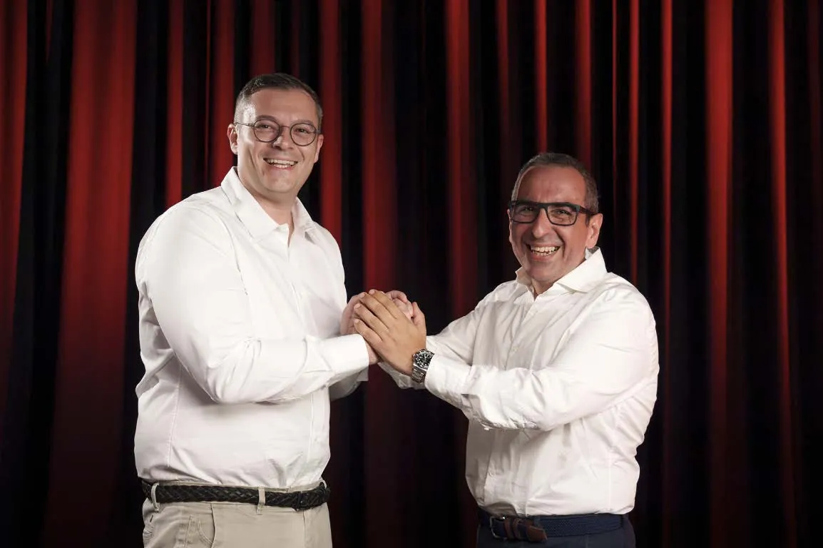 The two people leading the group shaking hands in front of a red stage curtain